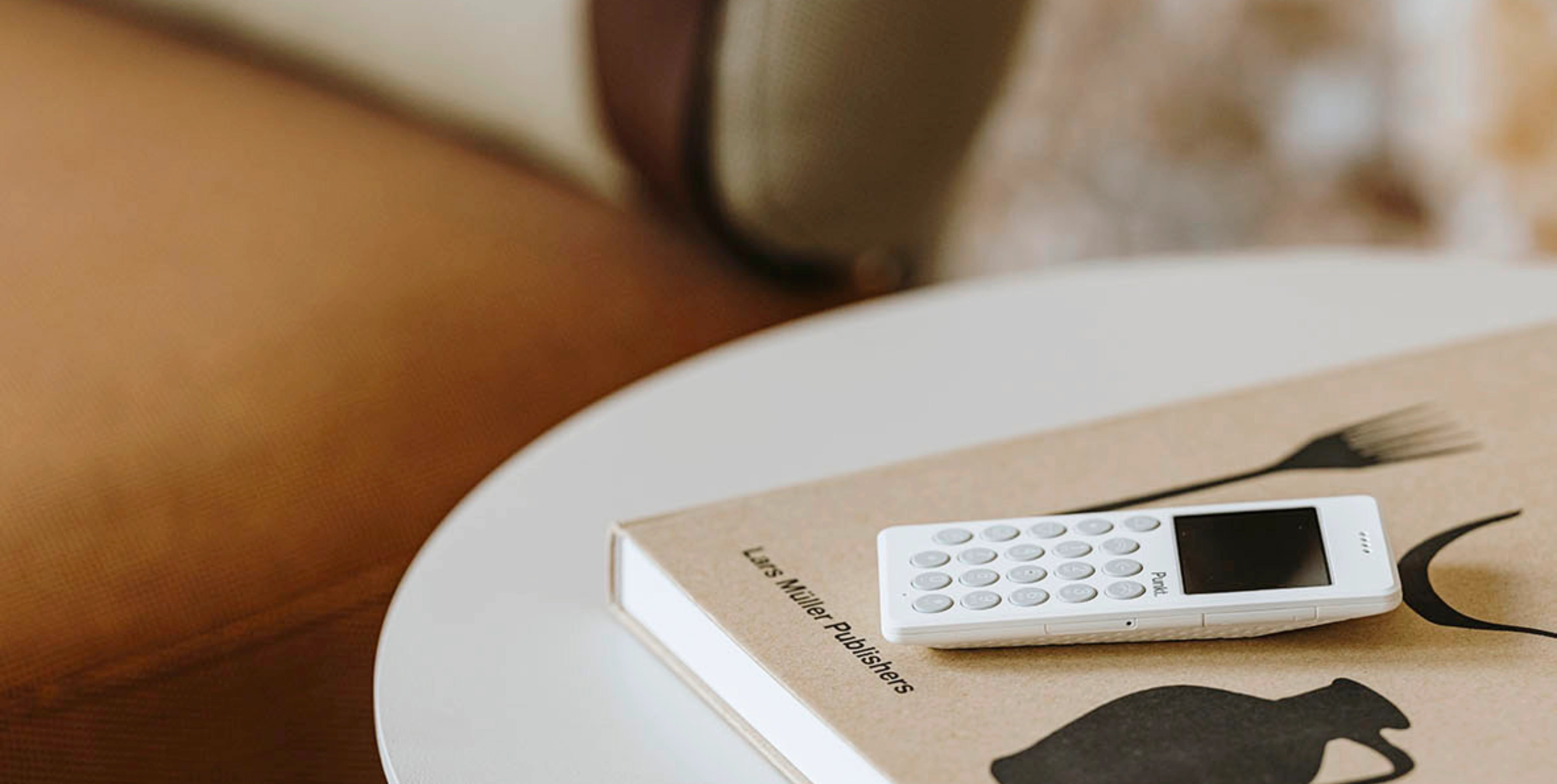 Punkt. MP01 mobile phone in white, resting on a Lars Müller Publishers book with minimalist graphics, atop a round table in a warm-toned interior.