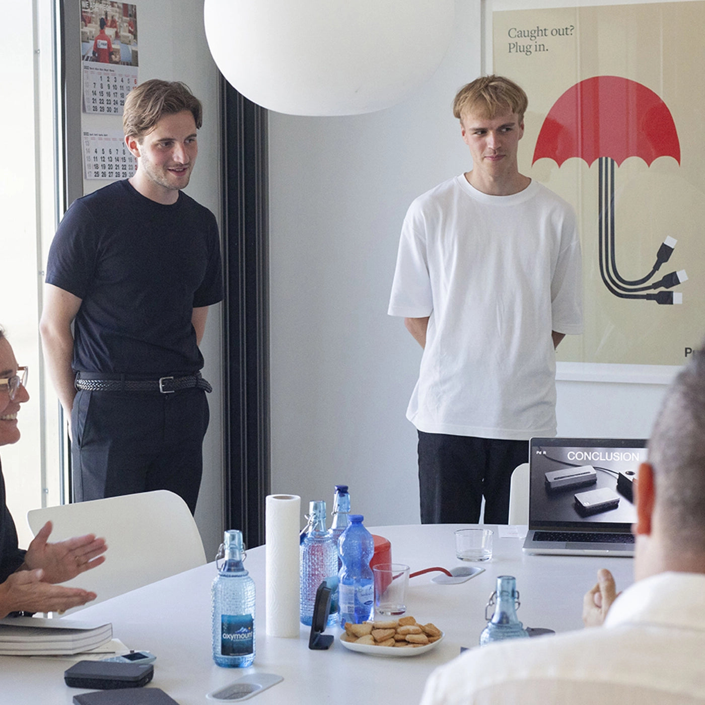 Four people in a meeting room, two standing and two seated at a table with laptops, bottles, and snacks; a presentation slide showing minimalist devices on screen.