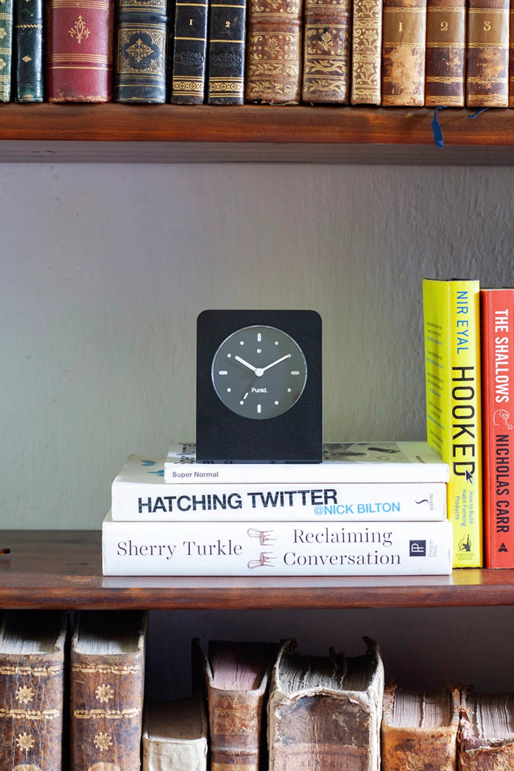 A minimalist Punkt. clock sits on a stack of modern books, framed by shelves of antique leather-bound volumes.