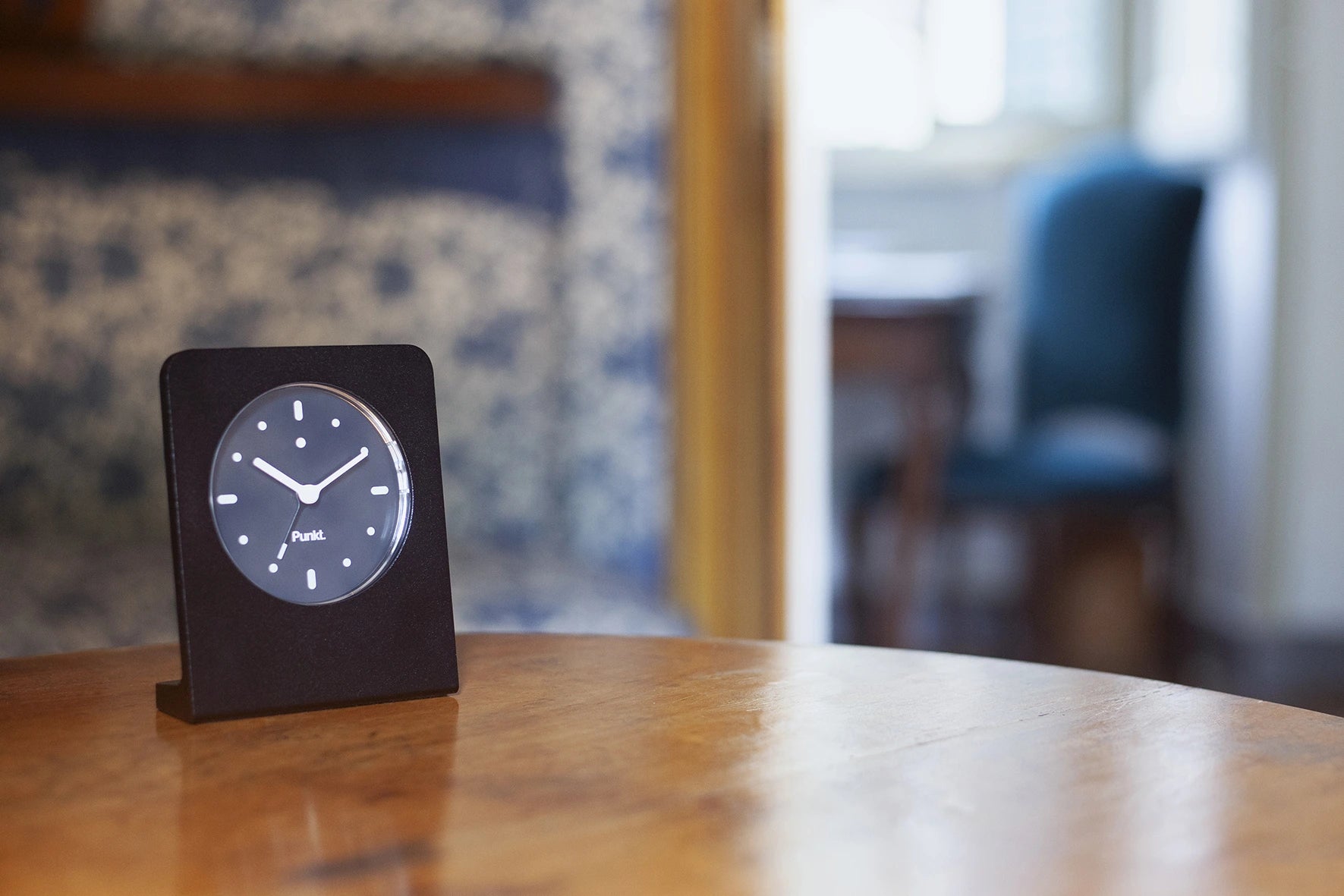Punkt AC02 clock on a wooden table in a traditional room with patterned wallpaper and soft lighting.