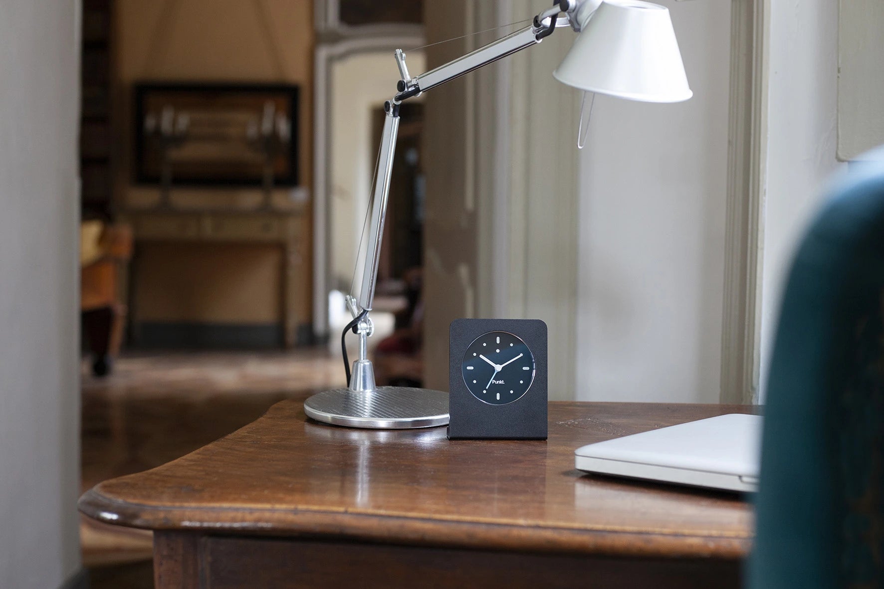 Punkt AC02 clock with blue second hand placed on a vintage desk beside a laptop and silver lamp, blending tech with tradition.