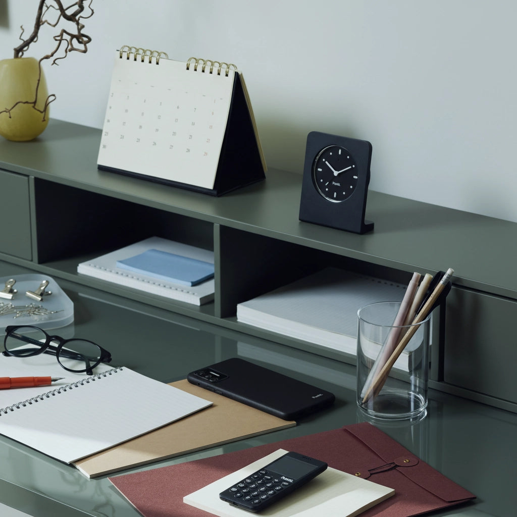 A neatly arranged desk featuring two phones, notebooks, a black desk clock, and a glass cup holding pencils and brushes.