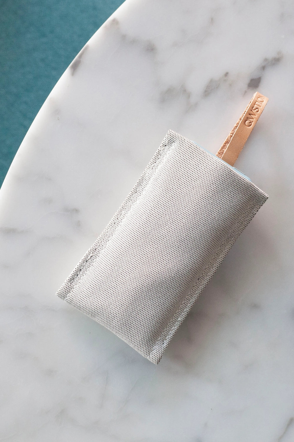 QWSTION fabric case for the Punkt. MP02 with leather loop, resting on a marble table; light blue phone partially visible.