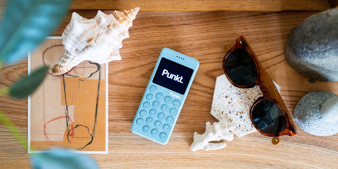A light blue Punkt. minimalist phone lies on a wooden tray surrounded by summer elements: a seashell, abstract artwork, tortoiseshell sunglasses, white coral, a terrazzo tile, and stacked stones, evoking a calm and natural digital detox setting.