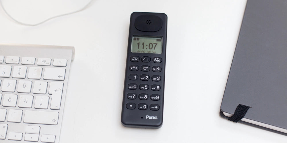 Black Punkt. DP01 cordless phone on a white desk, placed next to a keyboard and a closed black notebook.