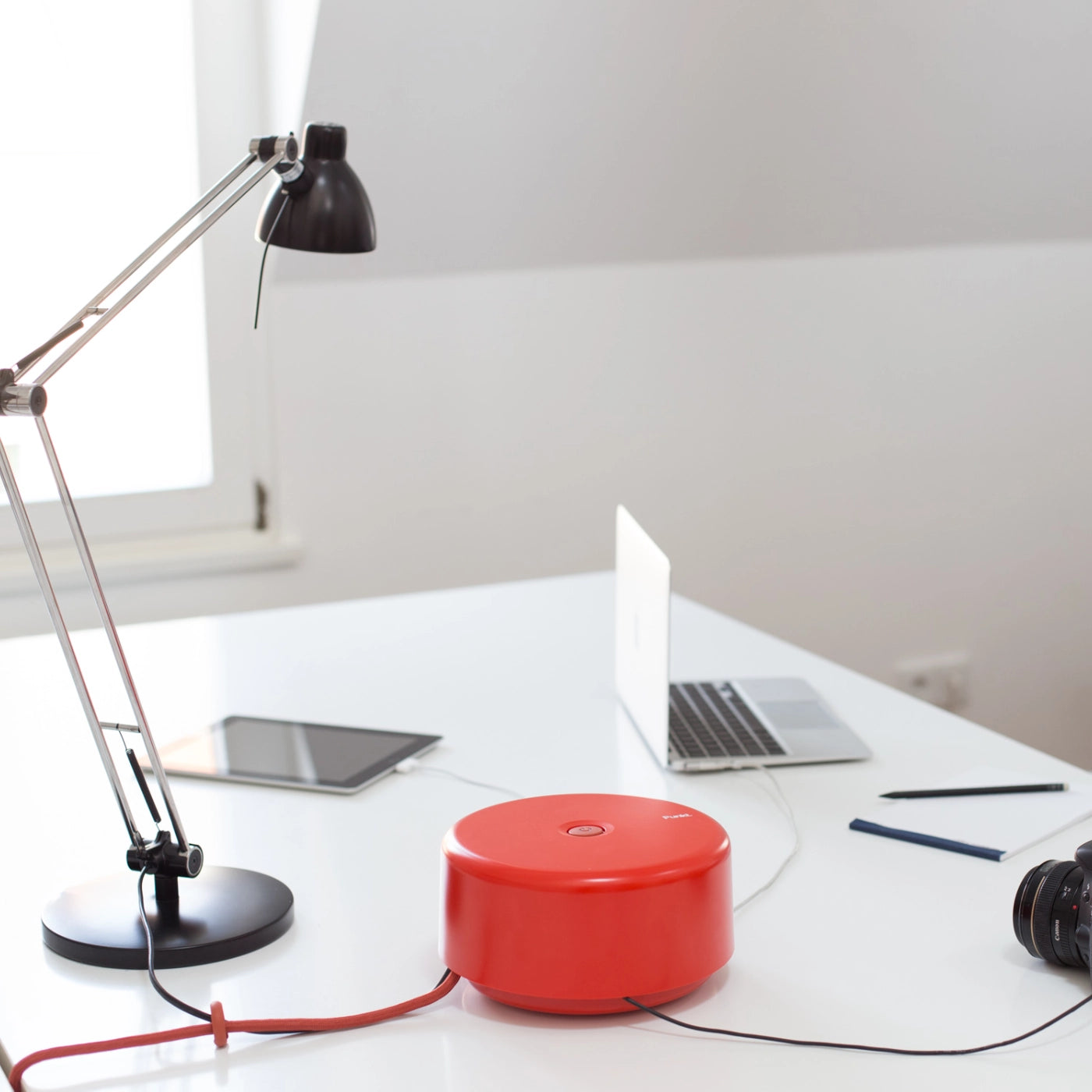 Red Punkt. ES01 on a white desk powering multiple devices, including a MacBook, tablet, and camera, in a bright room with a window lamp.
