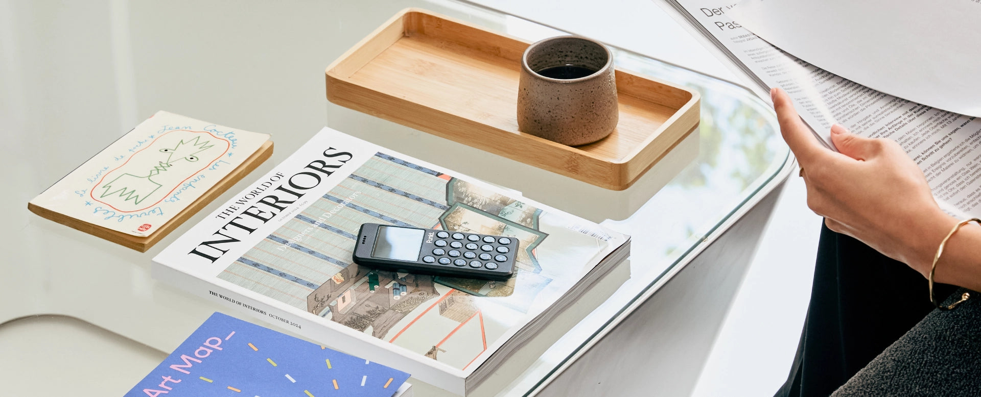 A minimalist tabletop scene with design magazines, including “The World of Interiors,” a Jean Cocteau book, and a blue art map booklet. A Punkt. mobile phone lies on the magazine, next to a wooden tray with a ceramic cup. A person is reading a printed document on the right side.