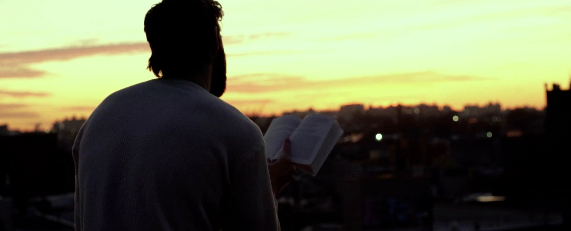 Man reading a book on a rooftop at sunset, overlooking a city skyline with warm orange and yellow tones.