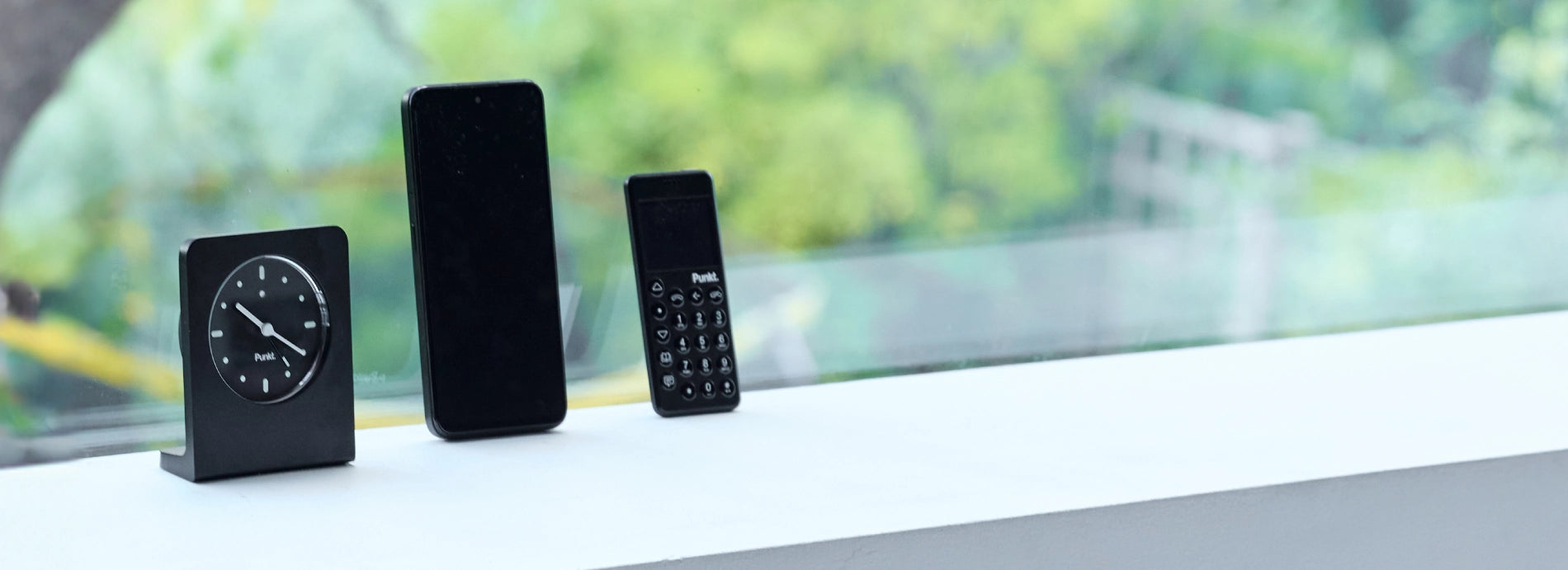 Minimalist display of a Punkt. clock, smartphone, and Punkt. phone arranged on a white ledge with a green, blurred outdoor background.