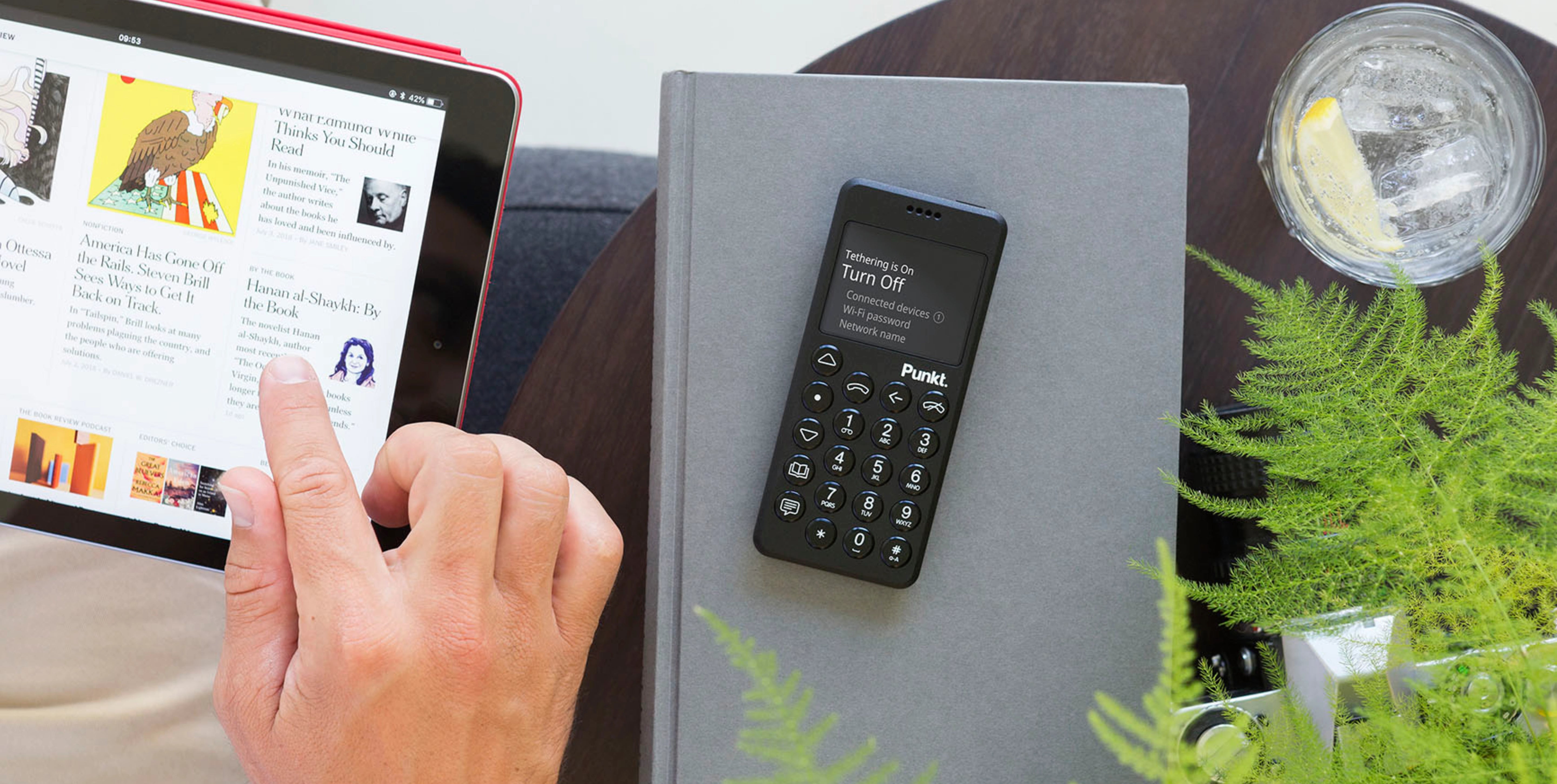 Person browsing a digital newspaper on a tablet, with a Punkt. MP02 phone placed on a closed book nearby, alongside a glass of iced lemon water and a green plant on a modern tabletop.