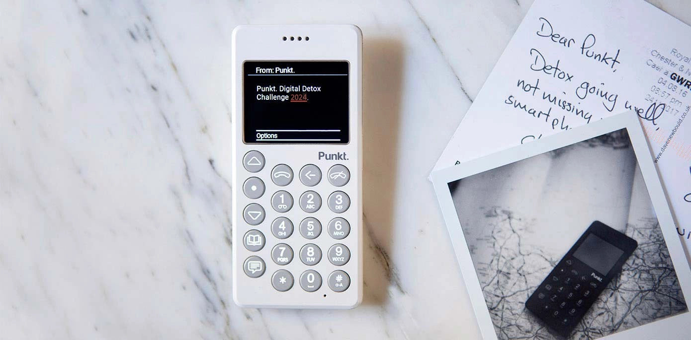 White Punkt. MP02 phone displaying a message about the Digital Detox Challenge 2024, placed on a white marble surface next to handwritten postcards and a black-and-white instant photo showing another Punkt. phone.