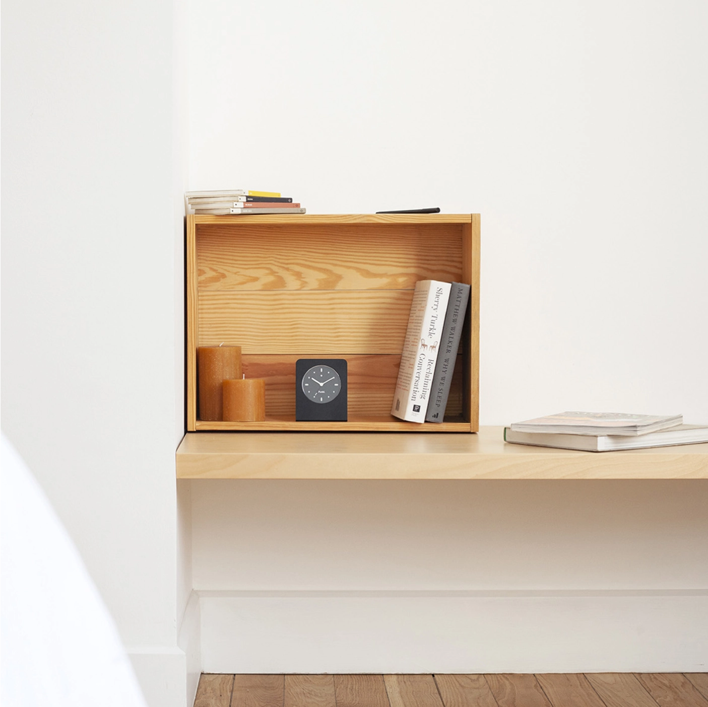 Punkt. AC01 black alarm clock displayed inside a wooden bookshelf next to candles and books in a calm reading nook.