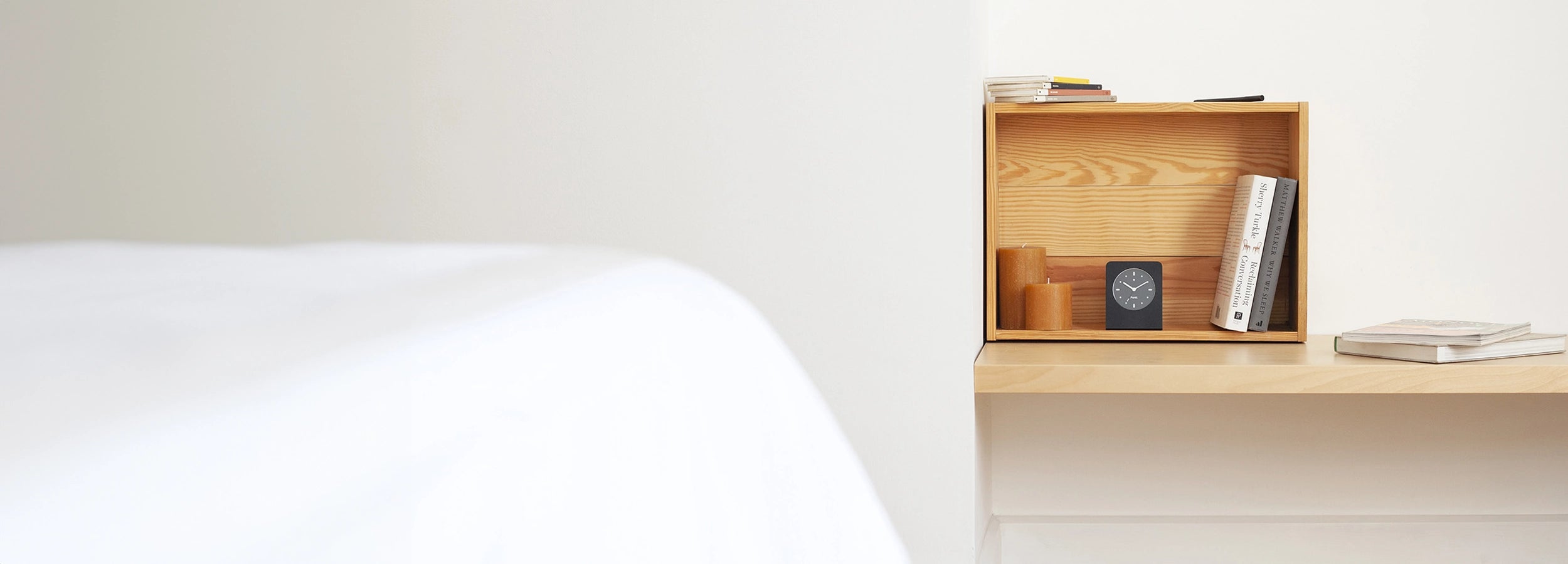  Minimalist wooden shelf with books, candles, and a small black alarm clock, placed beside a bed in a calm, neutral-toned bedroom.