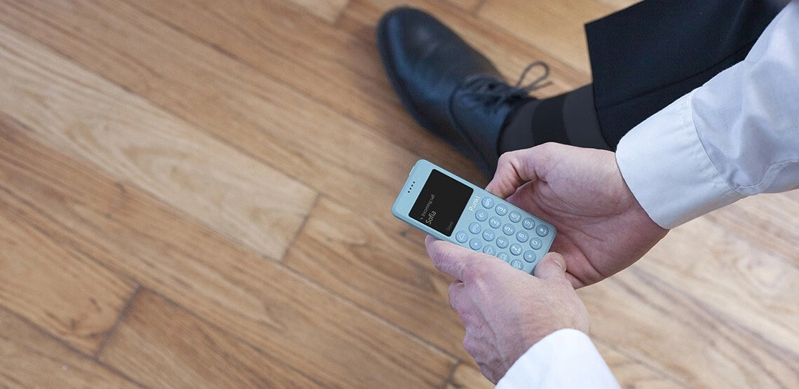 A person wearing a white shirt holds a compact light gray phone with physical buttons and a small screen, above a wooden floor.