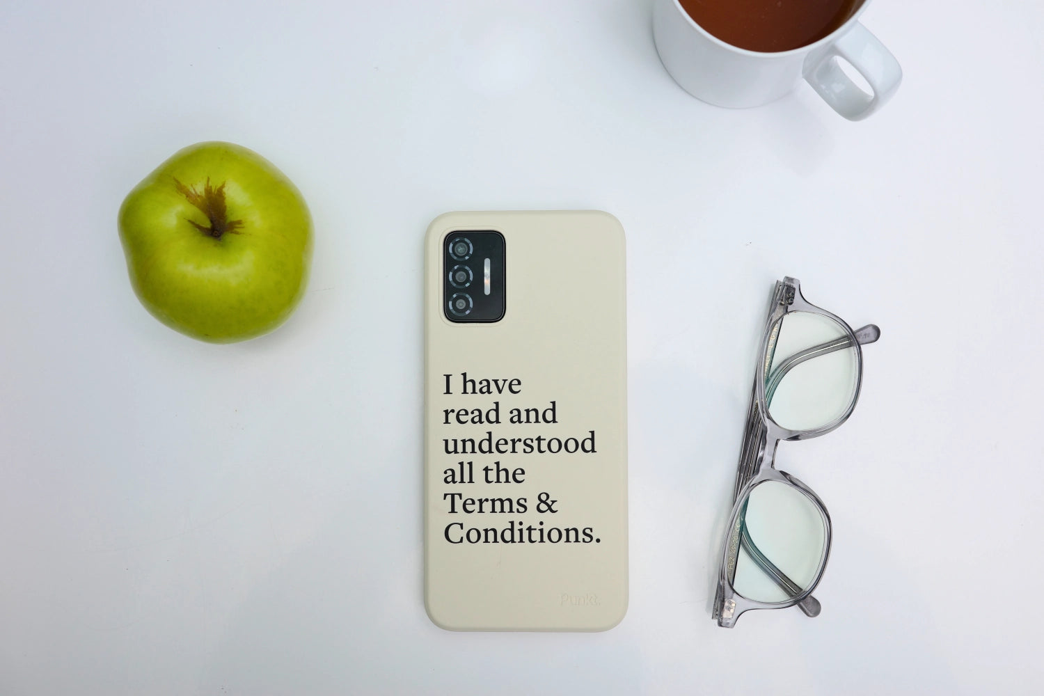 Flat lay of Punkt. MC02 smartphone in cream case with printed message, next to a green apple, glasses, and a cup of tea on a white table.
