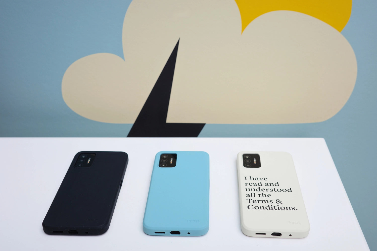 Three Punkt. MC02 smartphones placed on a white table with their bottom ports visible: black, light blue, and cream with printed message. Artistic background with abstract shapes.