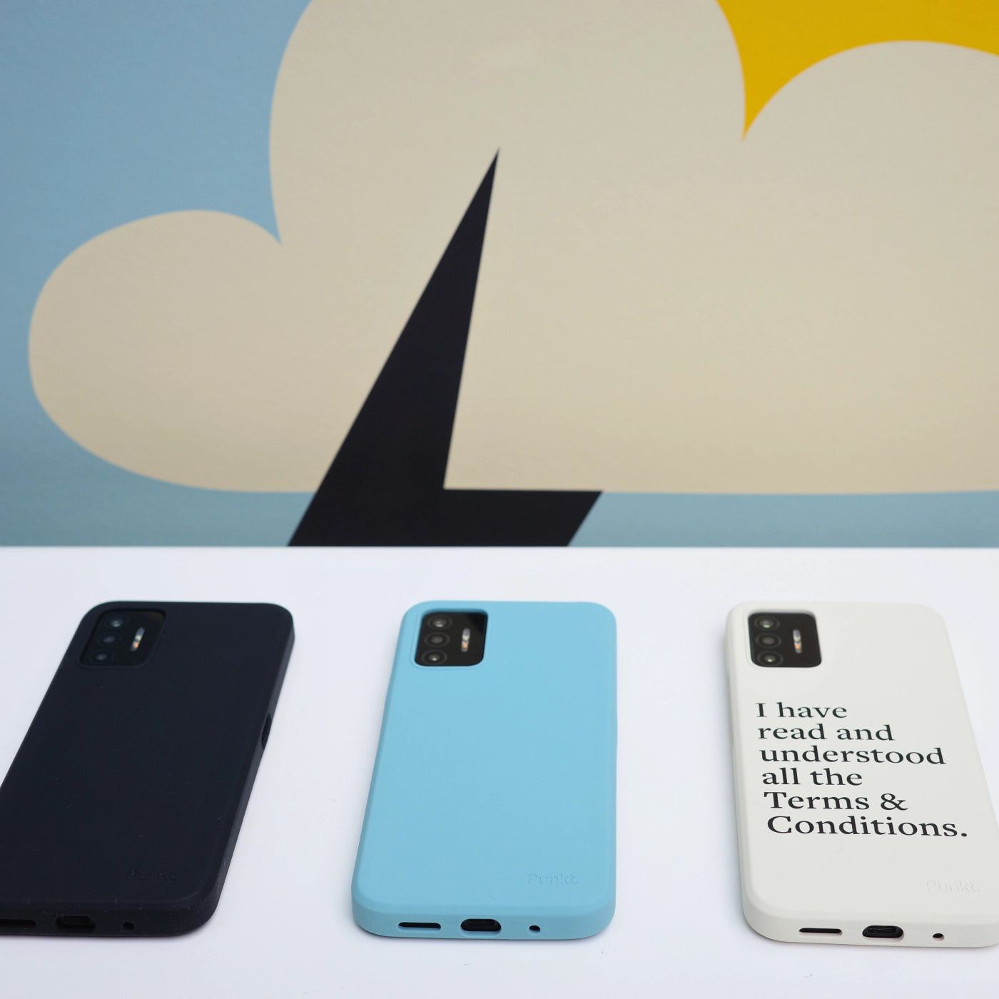 Three Punkt. MC02 smartphones placed on a white table with their bottom ports visible: black, light blue, and cream with printed message. Artistic background with abstract shapes.