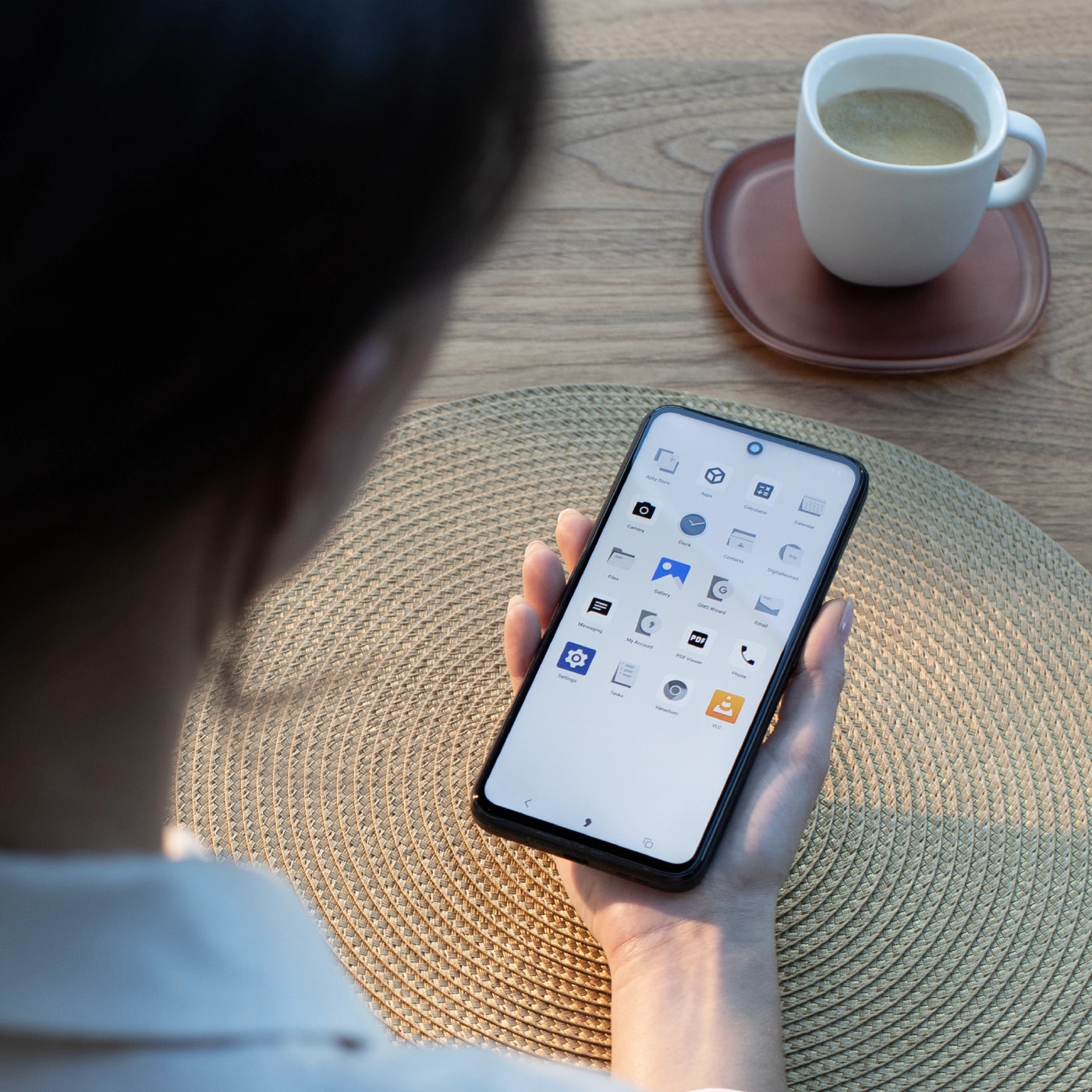 Hand holding smartphone displaying app icons on home screen, with coffee cup on saucer and woven placemat on wooden table.