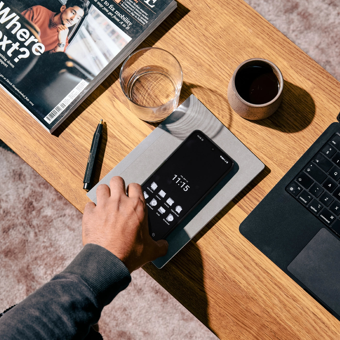 Hand using smartphone on wooden desk with laptop, coffee cup, water glass, pen, and magazine visible.