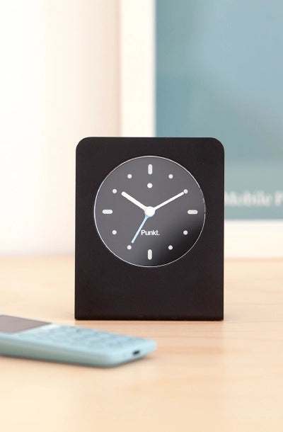 A sleek black Punkt. AC01 minimalist alarm clock displayed on a light wooden surface, with a soft blue mobile phone partially visible in the foreground and a blurred frame in the background.