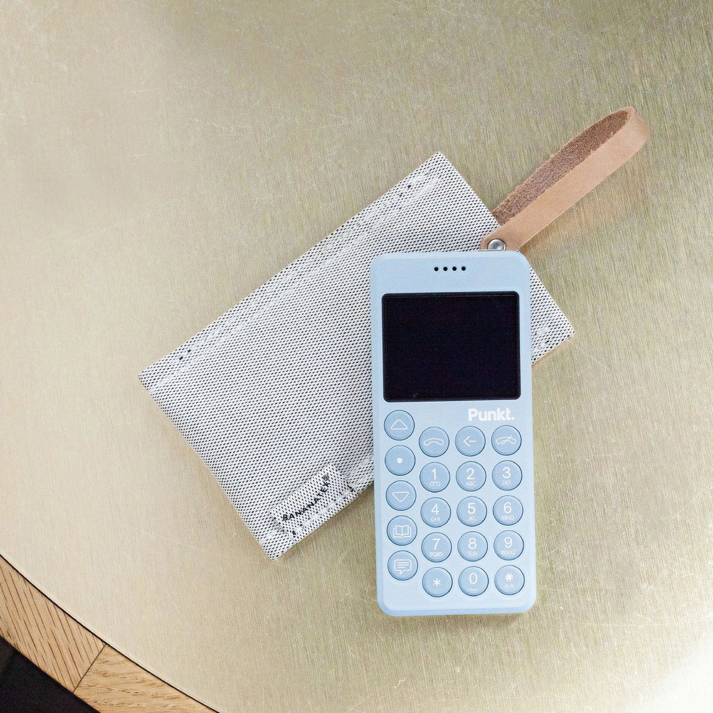 Top view of the light blue Punkt. MP02 and QWSTION grey sleeve with leather strap on a golden surface.