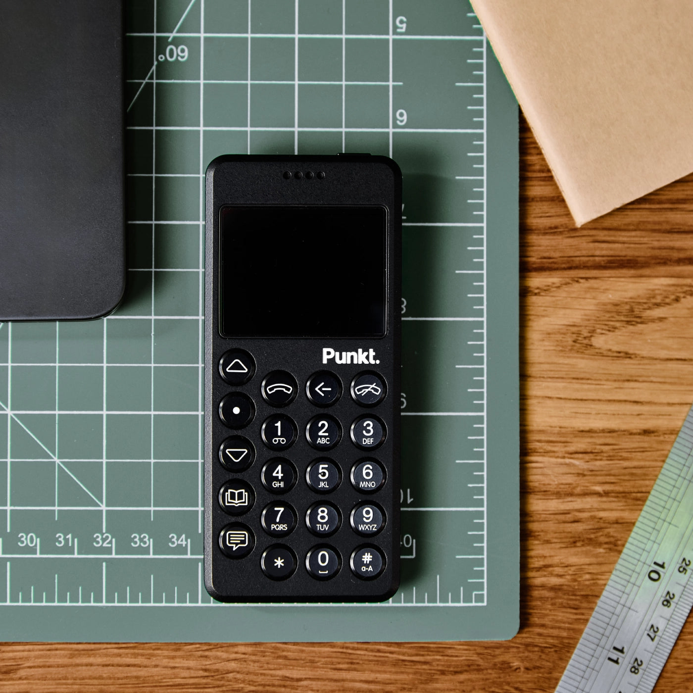 A black Punkt. MP02 phone placed neatly on a green cutting mat on a wooden desk.