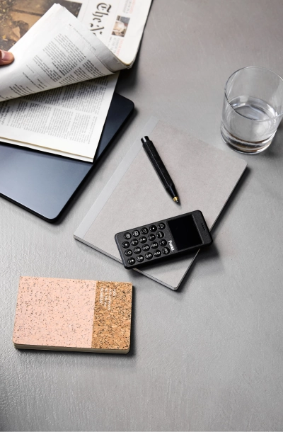 A black Punkt. MP02 placed on a grey desk, next to a notebook, pen, glass of water, and a cork-covered notepad.