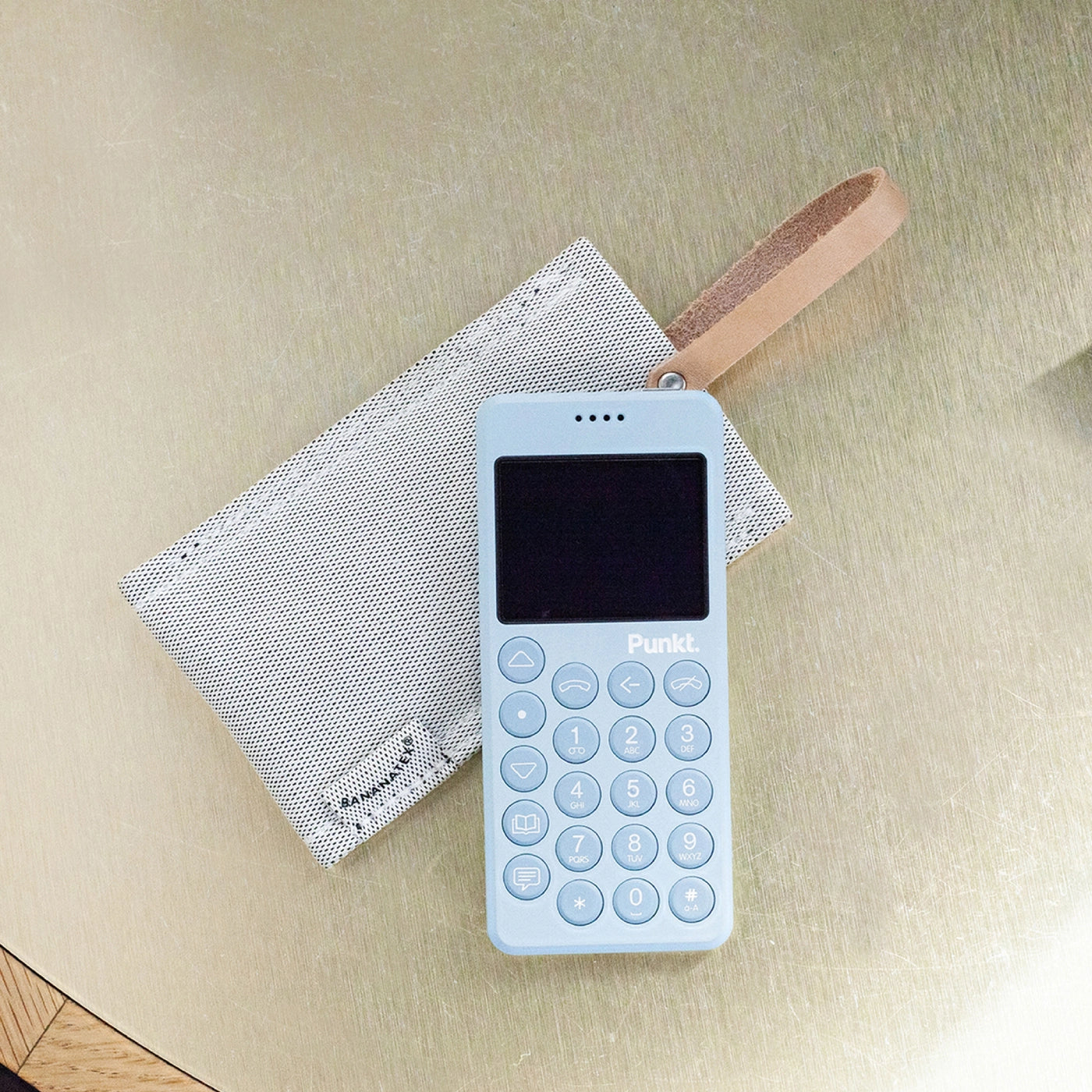 Light blue Punkt. MP02 phone resting on a QWSTION case with visible leather strap, on a metallic surface.