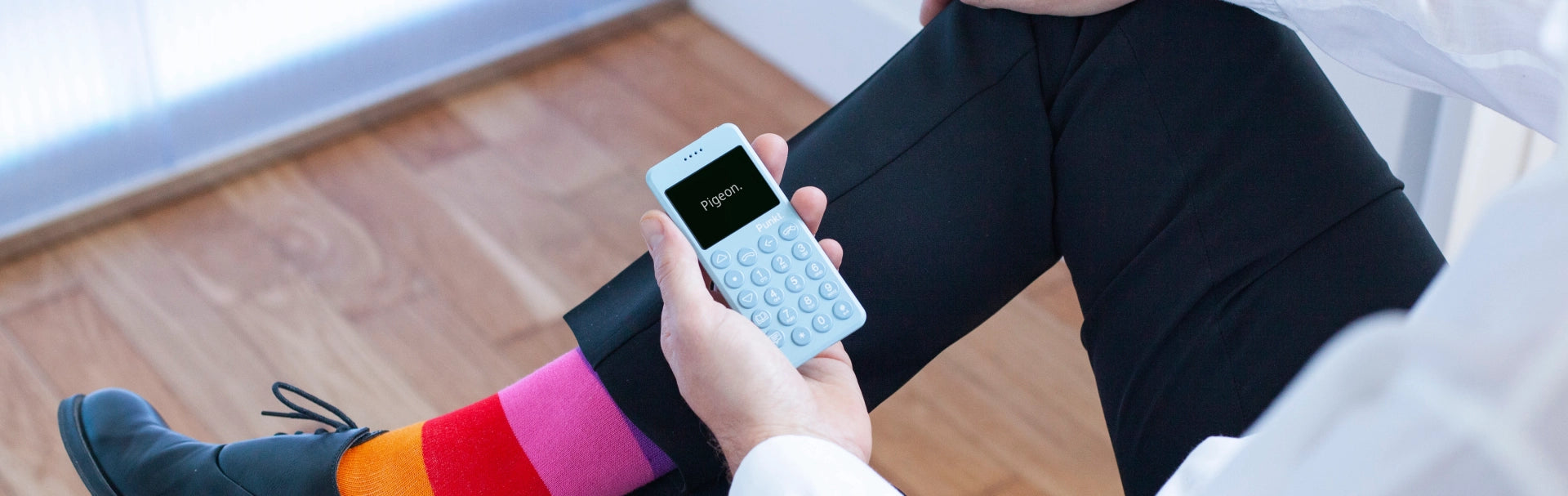 A man dressed in a white shirt, black pants, and colorful socks holds a light blue Punkt. phone displaying the word "Pigeon." on its screen while seated on a wooden floor.