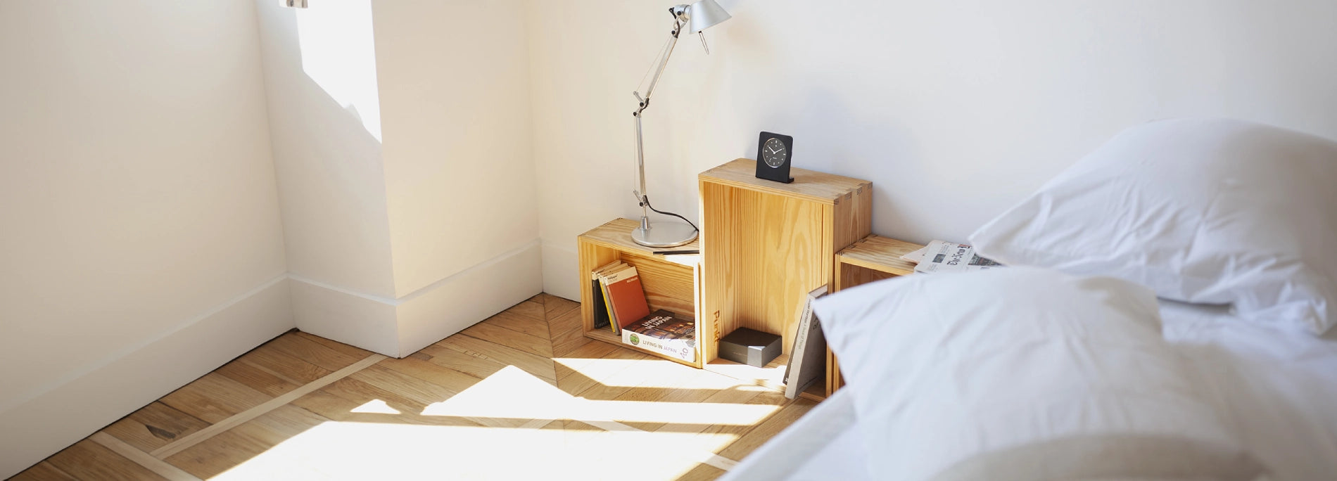 Camera da letto luminosa con comodino in cassette di legno, lampada e libri accanto a un letto bianco.