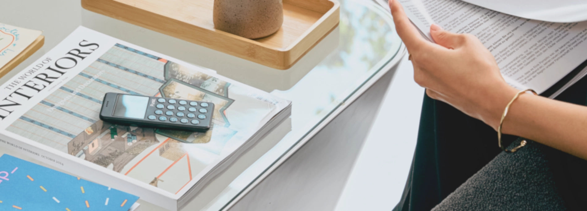 Close-up of a glass table with interior design magazines, a minimalist phone, and a person’s hand holding a document.