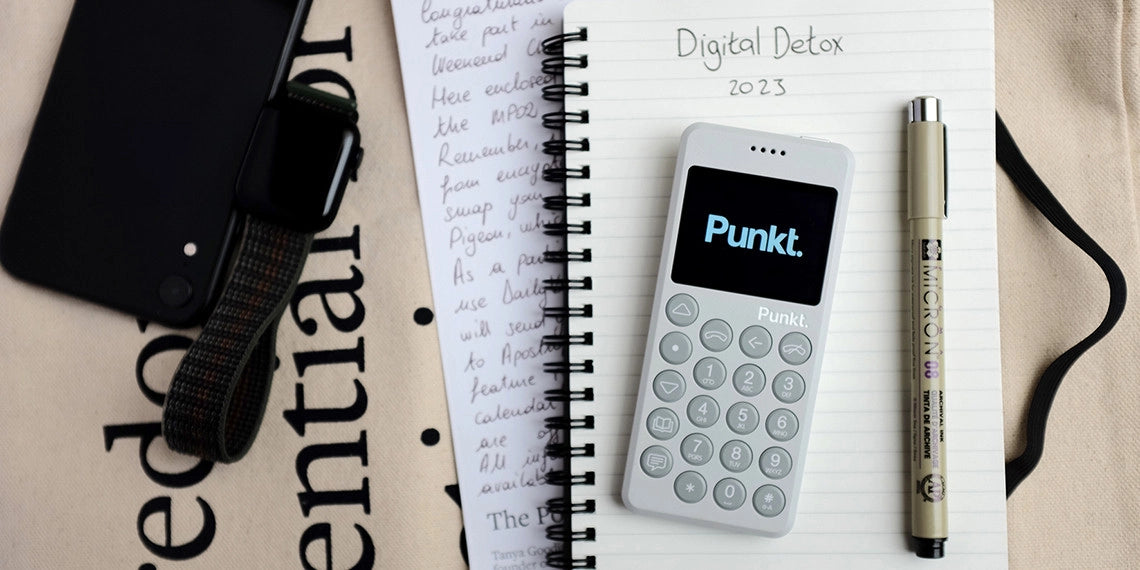 A light grey Punkt. phone rests on a spiral notebook labeled “Digital Detox 2023,” flanked by a beige Micron pen. Nearby are handwritten notes, a black smartwatch with a woven strap, and a black phone on a canvas tote bag printed with bold typography.