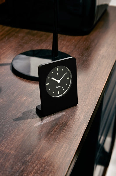 Minimalist black Punkt. AC02 with white hour markers placed on a polished wooden surface near a lamp base.