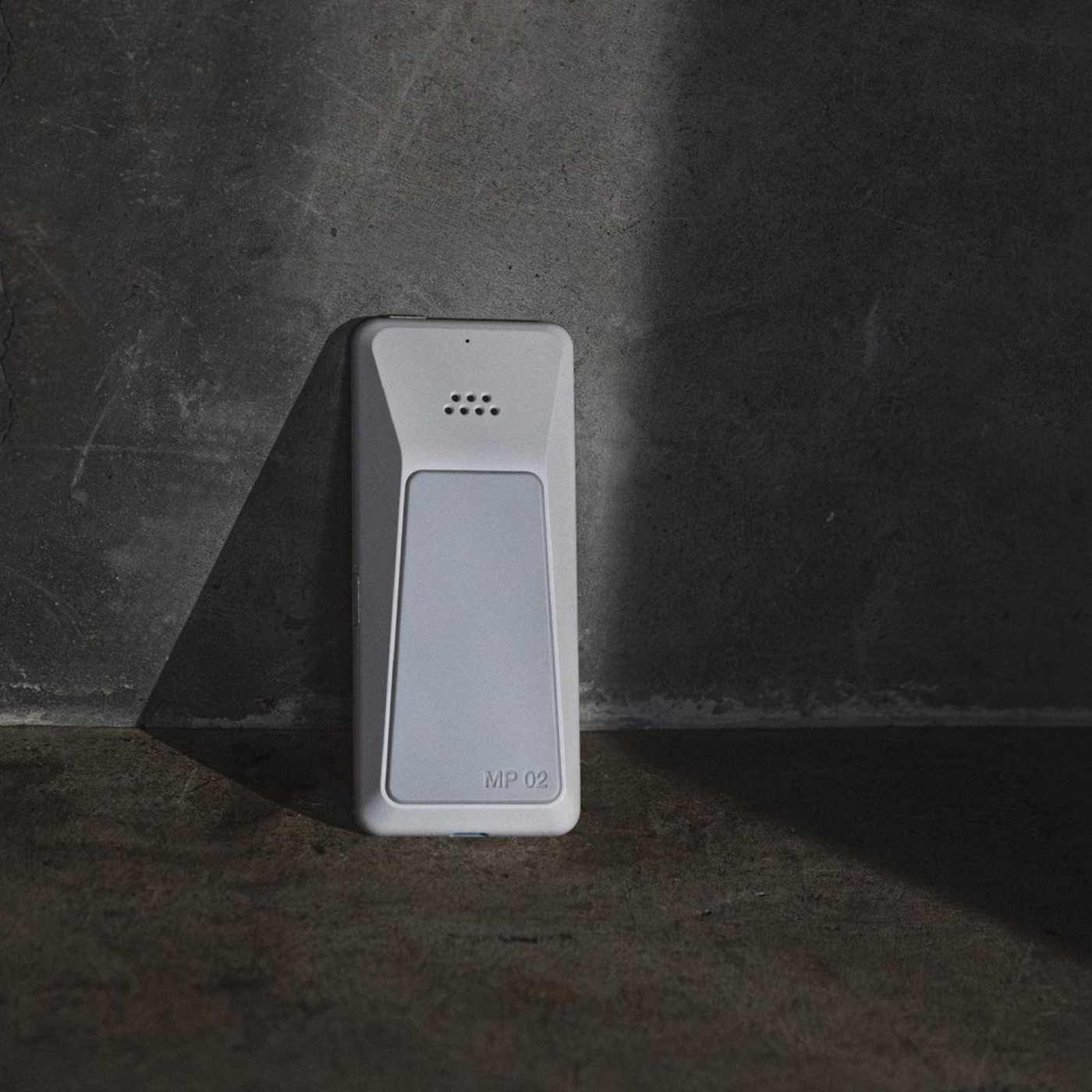 Minimalist product shot of the back of a light grey Punkt. MP02 phone, standing upright against a textured concrete wall with a beam of sunlight casting a diagonal shadow.