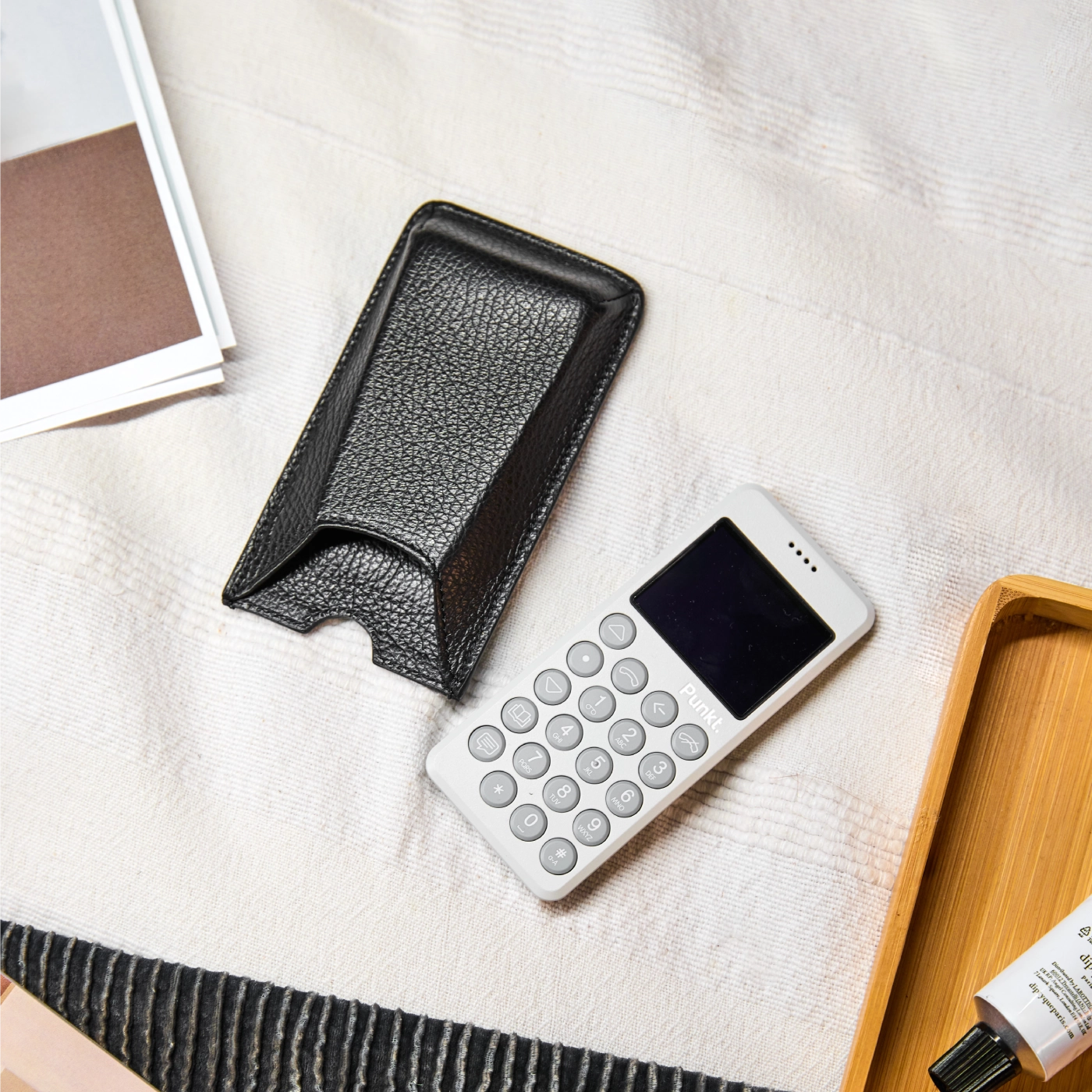White Punkt. MP02 mobile phone resting on a bed beside a black leather protective case, with books and a wooden tray visible in the background.