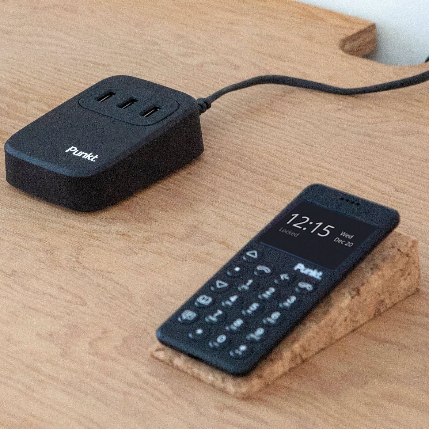 Punkt UC01 USB charger on a wooden desk next to an MP02 phone resting on a cork stand, illustrating a minimalistic and organized setup.