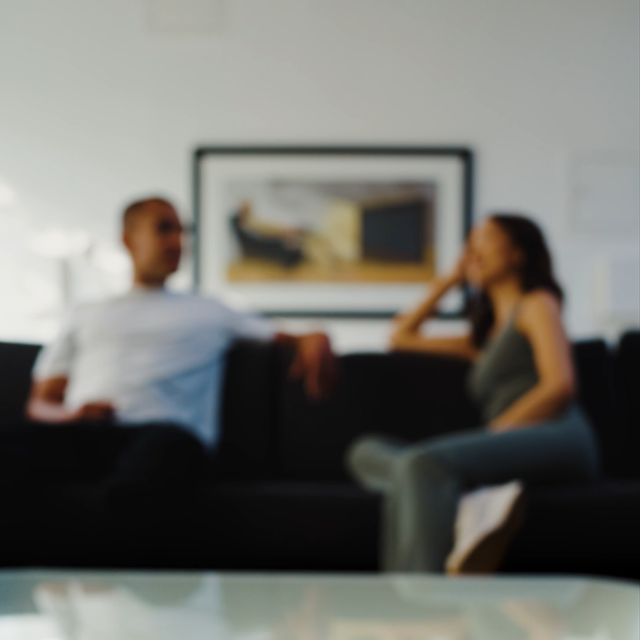 Intentionally blurred image of a man and woman sitting on a dark sofa, engaged in conversation in a modern, well-lit living room with a framed artwork in the background.