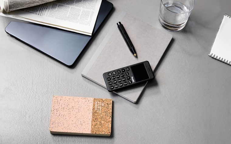 Black Punkt MP02 phone on a notebook with a pen, glass, and newspaper, symbolizing a privacy-focused life. 